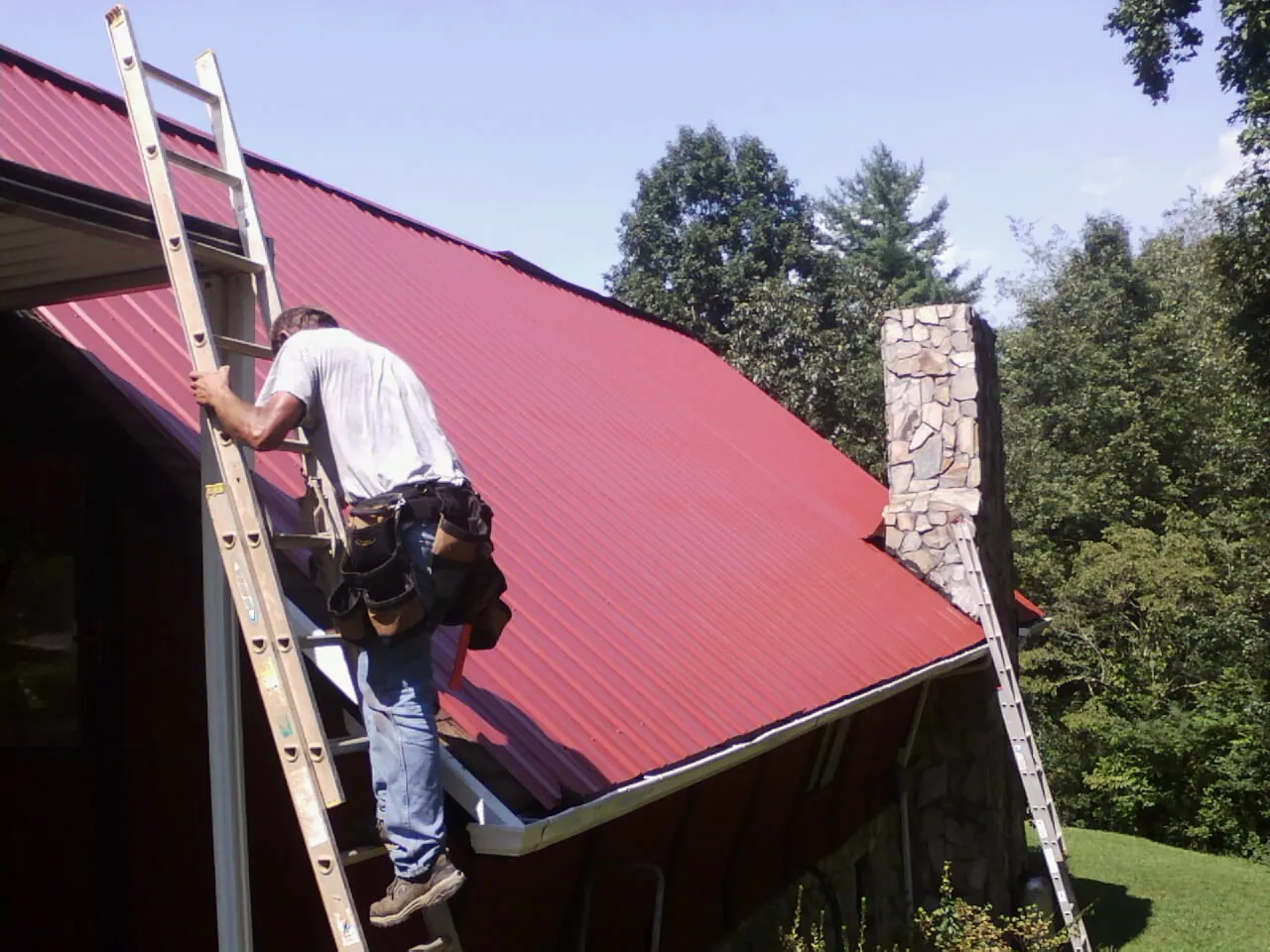 Roofer on a ladder beside a red metal roof and stone chimney — Soffit & Fascia Repair by Roofing Brothers in Hooker